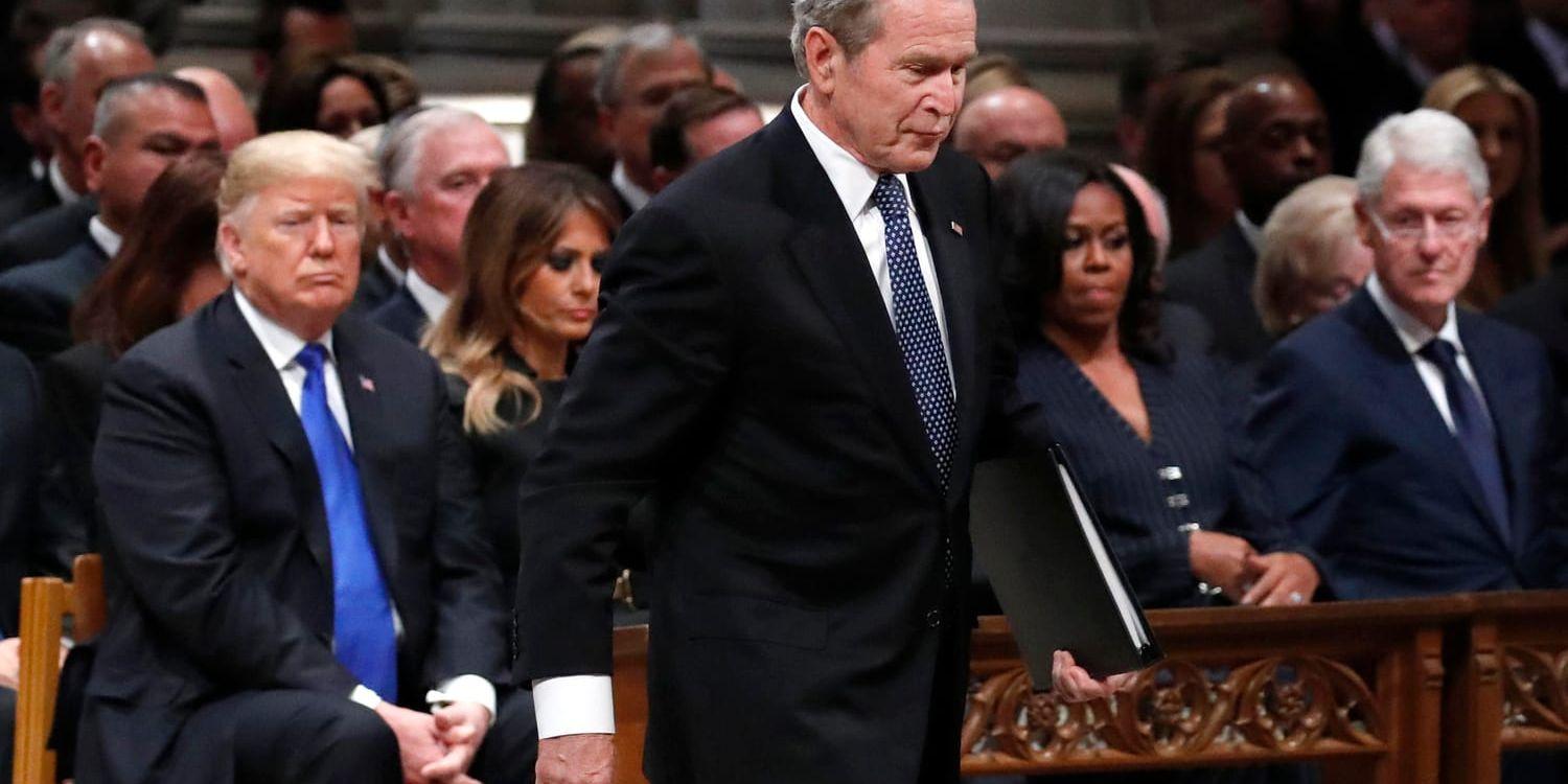Den tidigare presidenten George W Bush, son till George H W Bush, passerar USA:s nuvarande president Donald Trump vid ceremonin. I bakgrunden syns också USA:s första dam Melania Trump, USA:s tidigare första dam Michelle Obama, och den tidigare presidenten Bill Clinton.