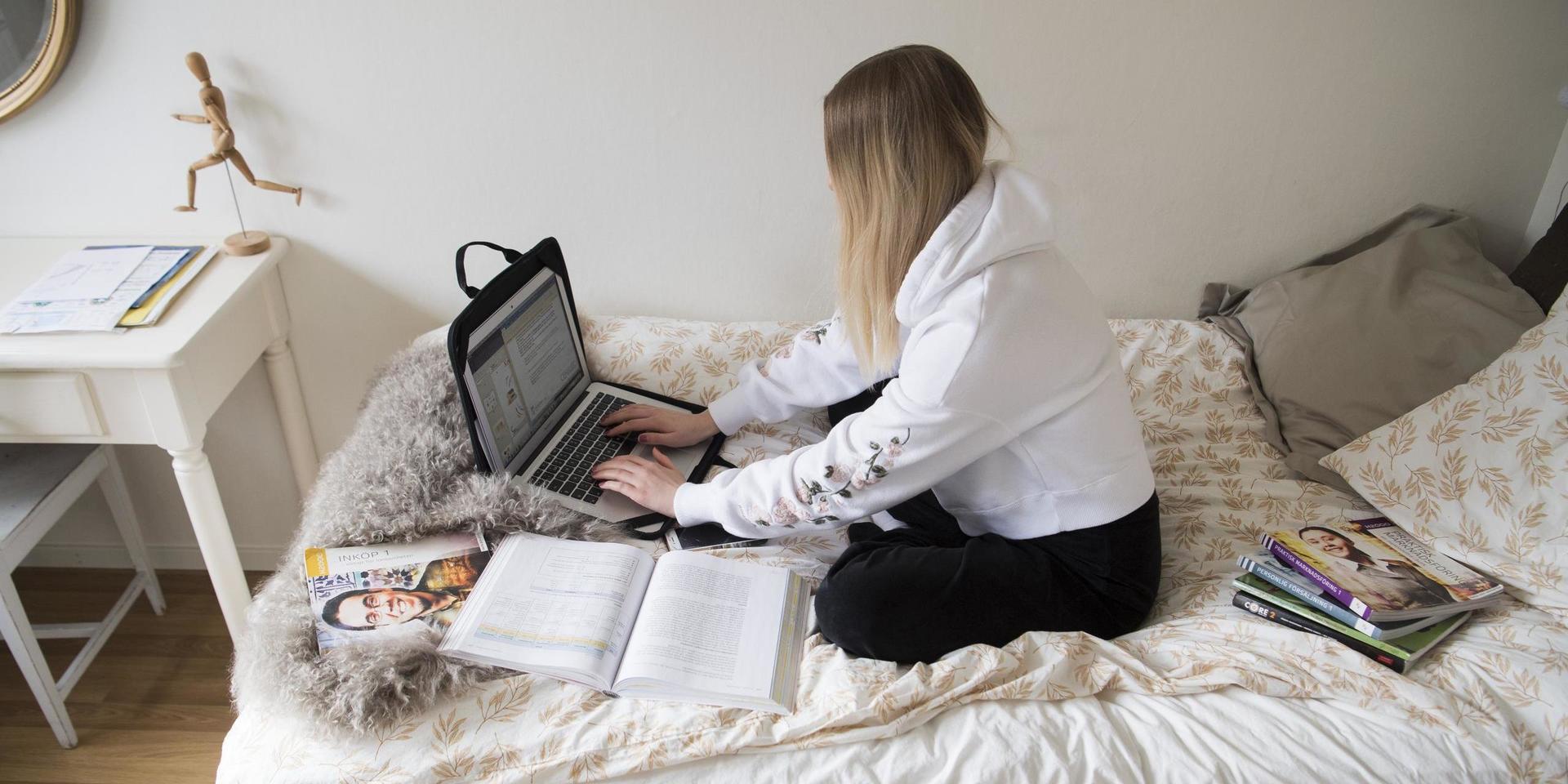 Gymnasieelev genomför hemundervisning via datorn, sittande i sängen.