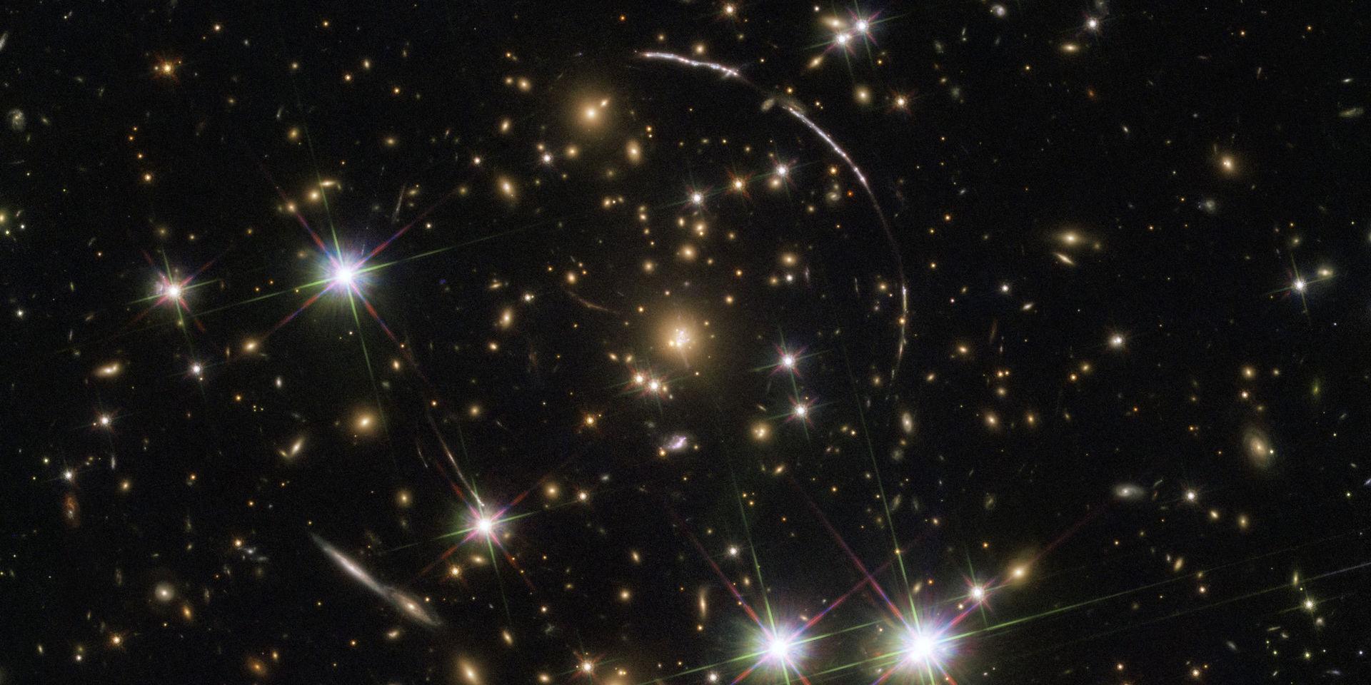 Bilden visar galaxklustret 4,6 miljarder ljusår bort. Längsmed syns fyra korta bågar – tre uppe till höger, en nere till vänster – från galaxen Sunburst arc, som ligger 11 miljarder ljusår bort.