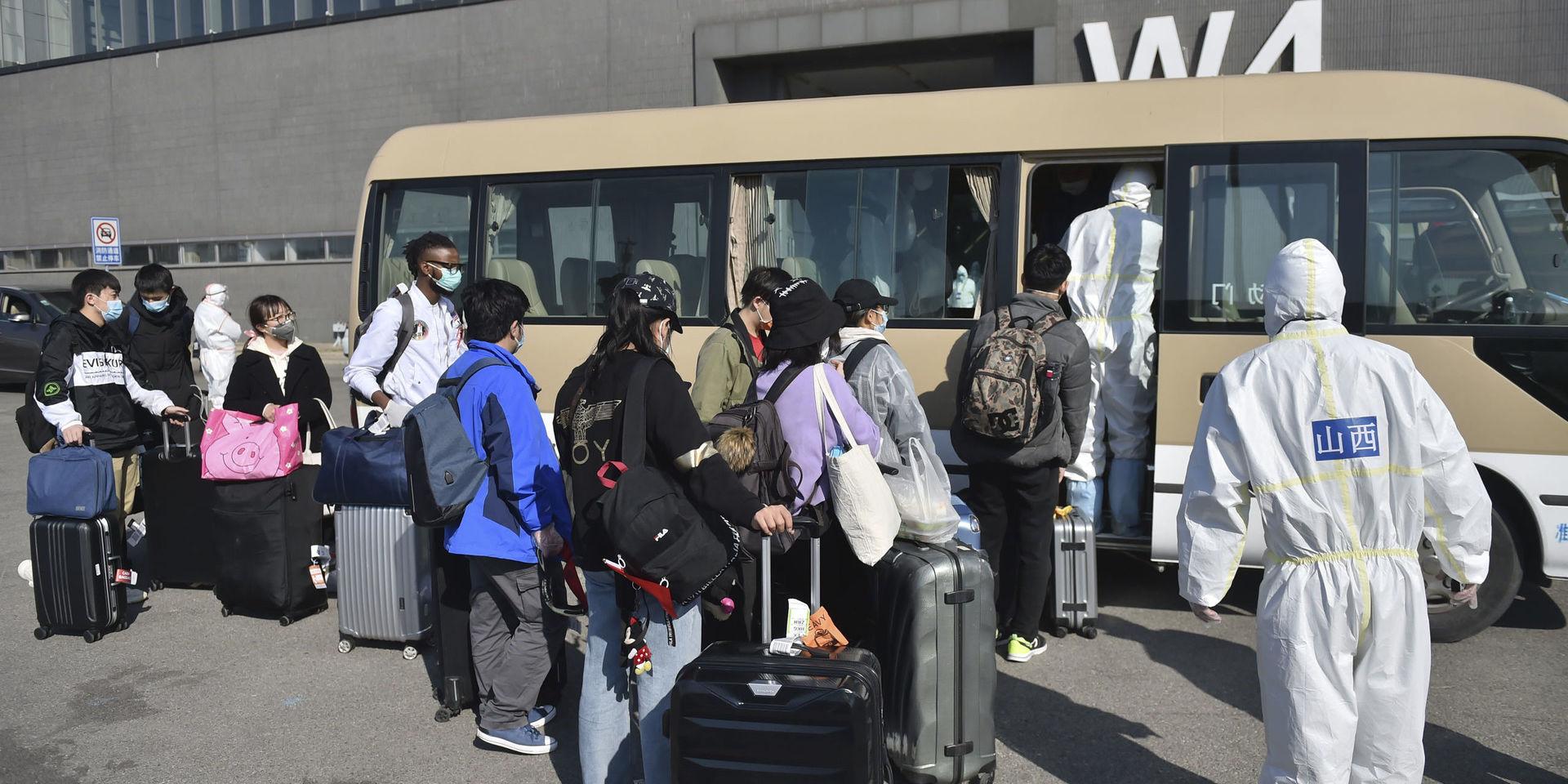 Smittskyddsarbetare ser på när resenärer kliver på en buss vid en utställningshall som gjorts om till en anläggning för att kontrollera personer som anlänt med flyg till Peking. 