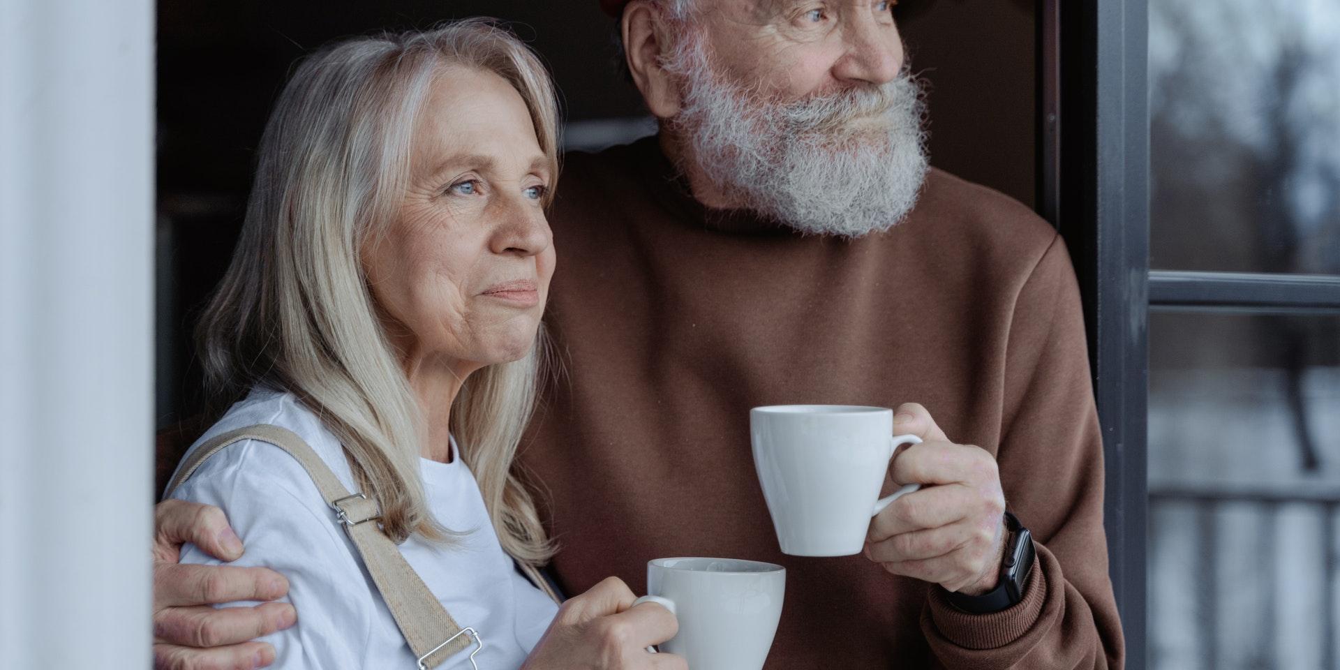 Äldre par tittar ut genom fönster, har kaffekoppar i händerna. pension pensionär äldre pensioner kärlek parrelation relation 